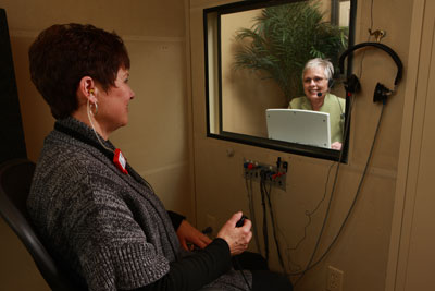 A woman sits in a soundproof booth wearing earphones for a hearing test, while a technician monitors and speaks to her through a glass window using a headset and microphone.