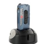 A small blue handheld device with buttons and a display screen is placed upright in a black charging dock, shown against a white background.