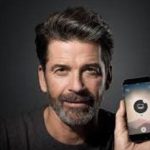 A middle-aged man with dark hair and a beard is smiling and holding up a smartphone displaying an app with a circular icon on the screen, against a dark background.