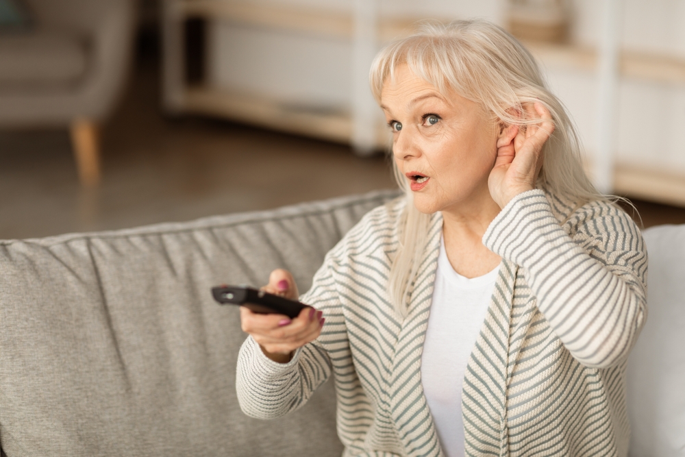 Older woman sitting on a couch, holding a TV remote in one hand and cupping her ear with the other, appearing to have difficulty hearing the television.