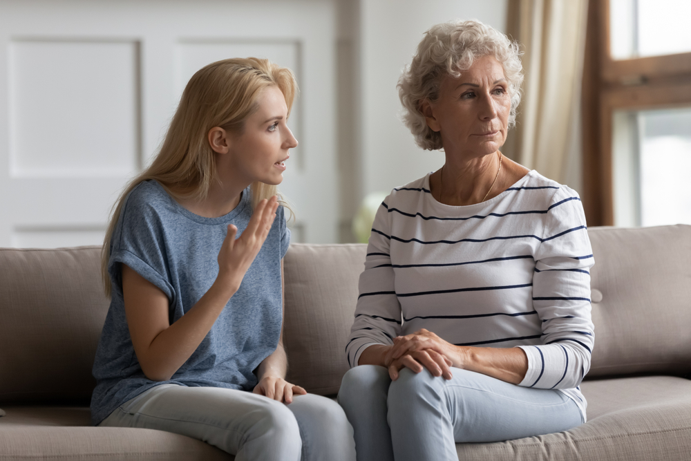 A younger woman is speaking emotionally to an older woman, who looks away with a serious expression. They are sitting on a couch in a bright living room, suggesting tension or disagreement between them.