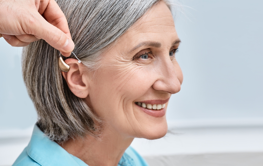 Smiling older woman with gray hair has a hearing aid placed behind her ear by another persons hand, illustrating use of hearing assistance device.