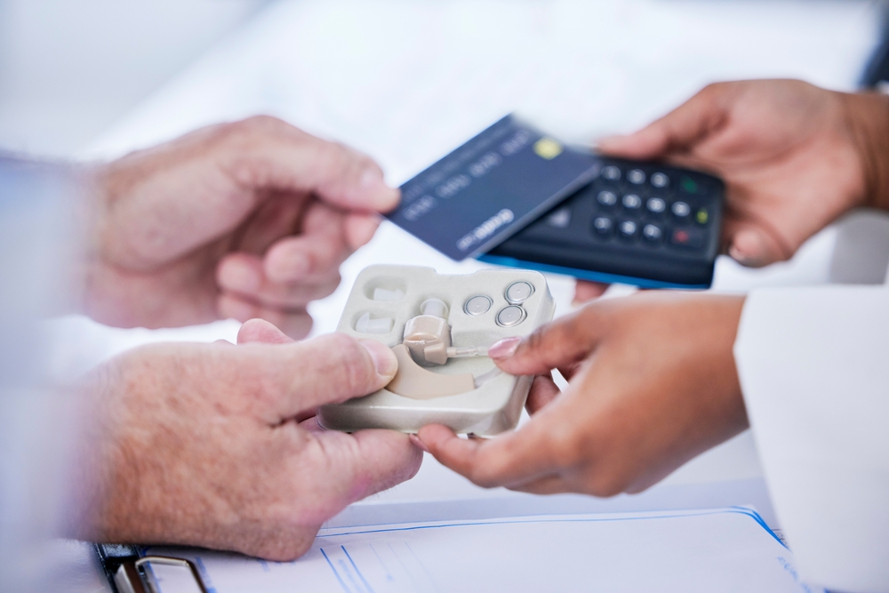 A person hands a credit card to another person holding a card reader, while also receiving a hearing aid device in packaging. A document and pen are visible on the table below.