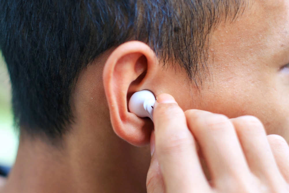 A close-up of a person inserting a white wireless earbud into their right ear, with their hand visible adjusting the earbud.