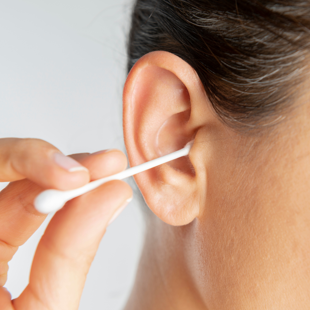 A person holds a cotton swab and gently cleans the outer part of their ear. The background is neutral and the image focuses on the ear and the swab.