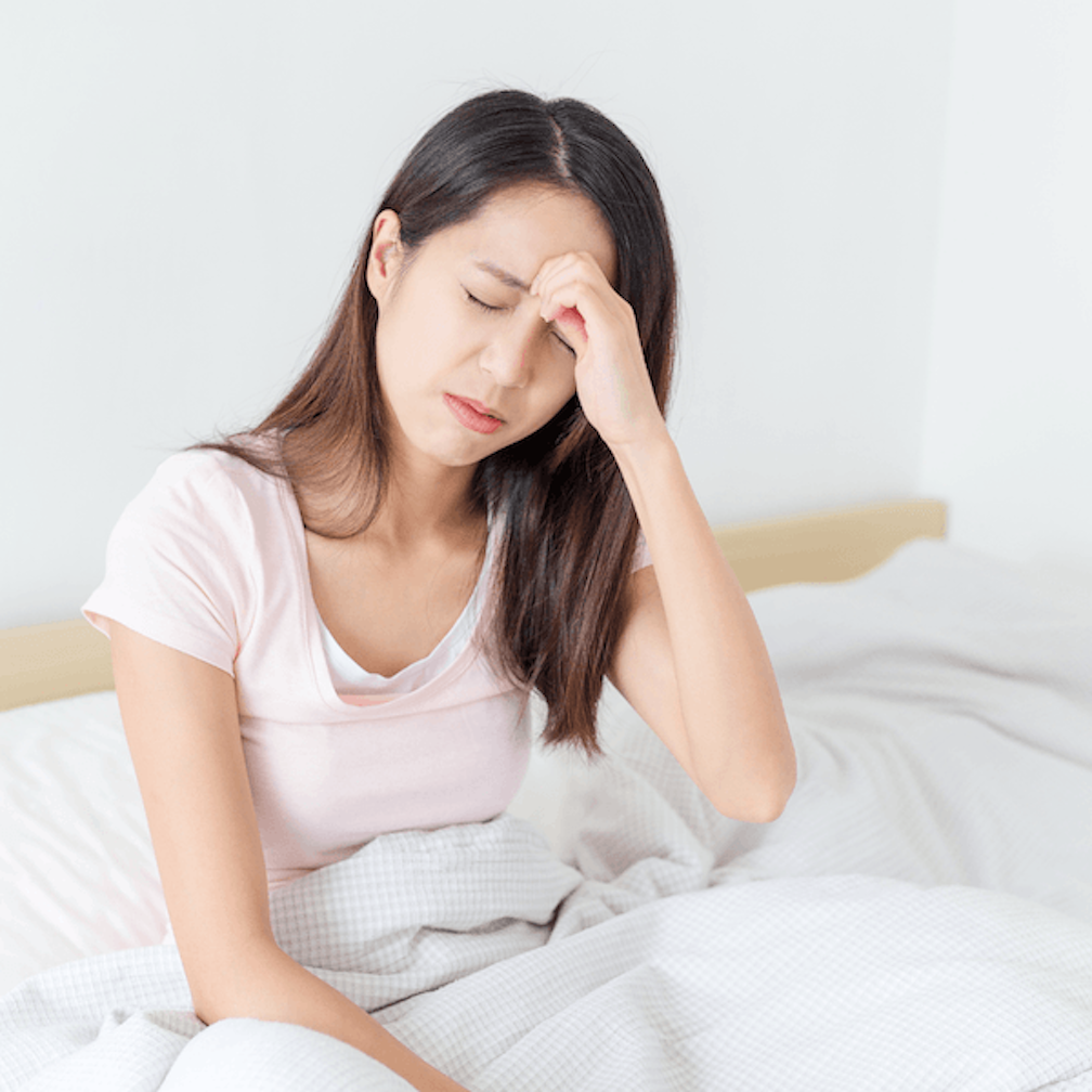A woman sits on a bed with her eyes closed, holding her forehead with one hand, appearing to have a headache or feel unwell.