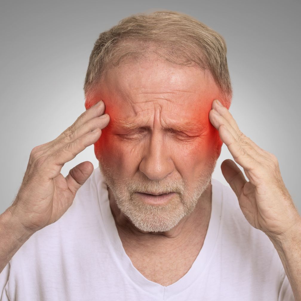 An older man with gray hair holds his head with both hands and closes his eyes in pain. Red areas on his temples indicate headache or migraine discomfort.