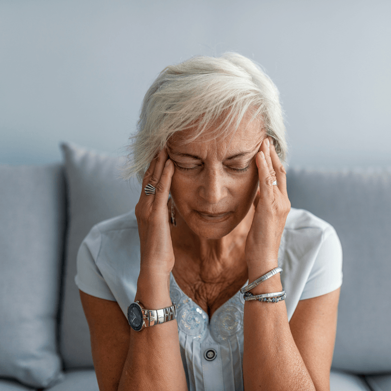 An older woman with short gray hair sits on a sofa, eyes closed, pressing her temples with her fingers as if experiencing a headache or stress. She wears a light blue blouse and silver jewelry.