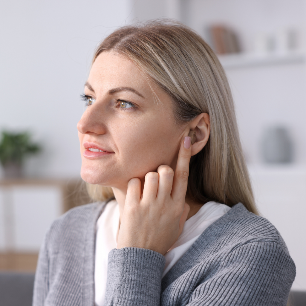 A woman with long blonde hair touches her ear with her finger, appearing thoughtful. She is wearing a gray sweater and is indoors in a softly lit room with blurred shelves in the background.