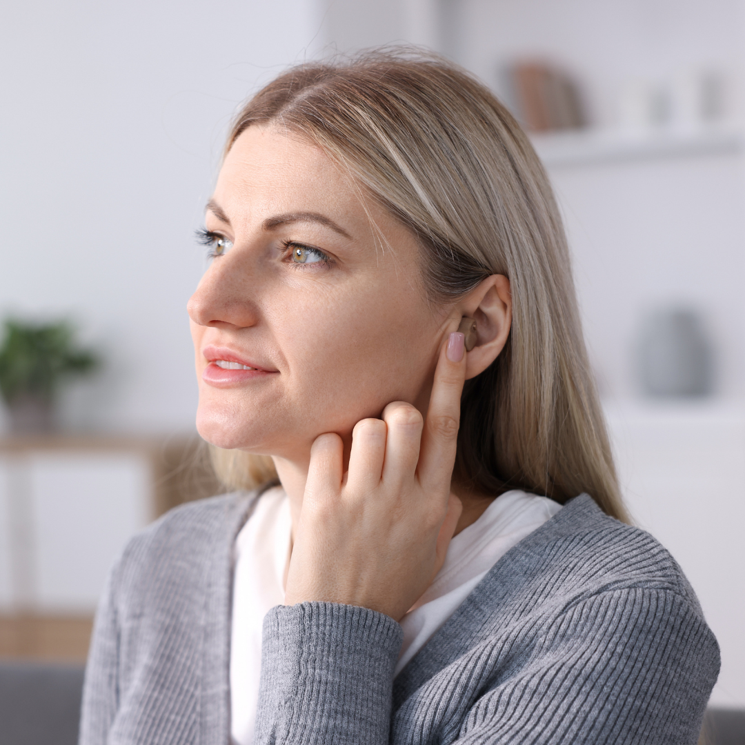 A woman with long blonde hair touches her ear with her finger, appearing thoughtful. She is wearing a gray sweater and is indoors in a softly lit room with blurred shelves in the background.