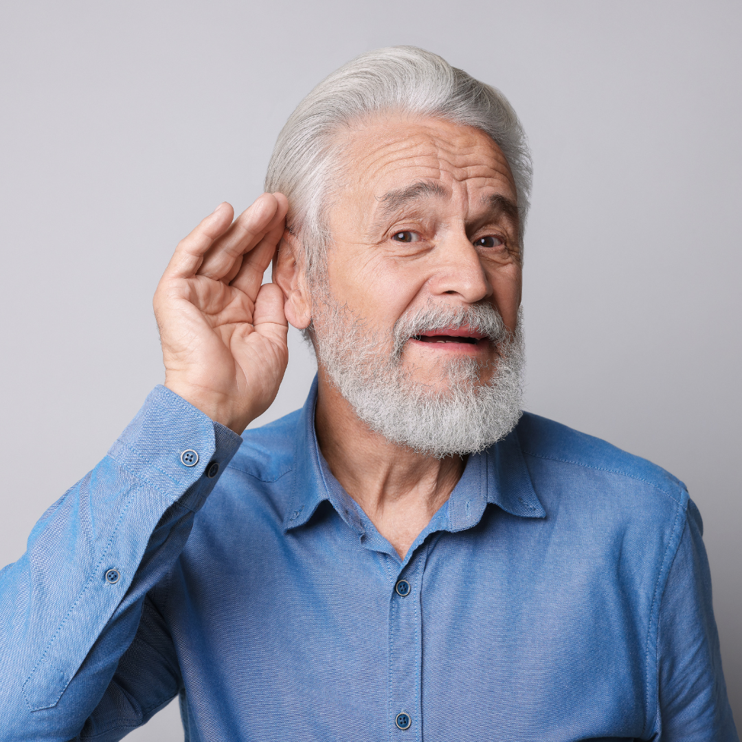 Older man with gray hair and beard wearing a blue shirt, holding his hand to his ear in a listening gesture, looking at the camera with a slight smile. Plain light background.